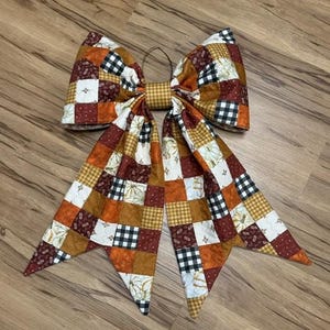 May include: A large decorative bow featuring a patchwork design of fabric squares in shades of orange, brown, white, and black. The pattern includes buffalo plaid, solid colors, and pumpkin prints, with a central knot and two long, pointed tails.
