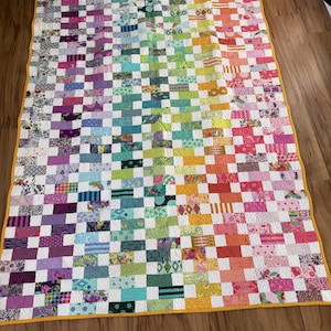 Könnte beinhalten: Eine handgefertigte Steppdecke mit einem Regenbogen-Farbverlauf. Die Steppdecke hat ein Patchworkmuster aus verschiedenen Stoffen in Lila, Blau, Grün, Gelb, Orange und Rot. Die Steppdecke ist mit einem gelben Stoff eingefasst.