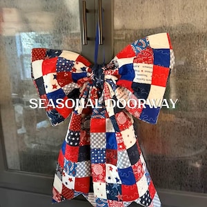 May include: A large decorative bow made of quilted fabric featuring a patchwork pattern in red, white, and blue. The fabric displays various star motifs, geometric shapes, and text. A blue ribbon loop is attached for hanging. The text SEASONAL DOORWAY is visible.
