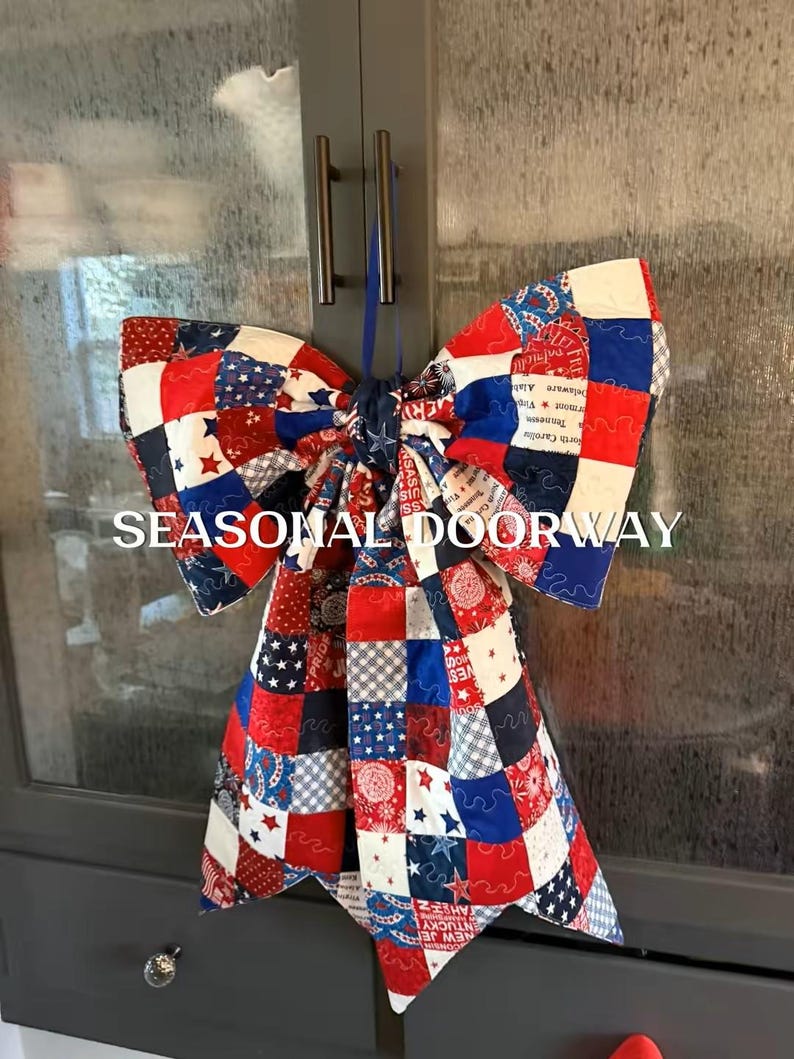 May include: A large decorative bow made of quilted fabric featuring a patchwork pattern in red, white, and blue. The fabric displays various star motifs, geometric shapes, and text. A blue ribbon loop is attached for hanging. The text SEASONAL DOORWAY is visible.