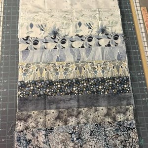 May include: A collection of twelve rectangular fabric strips arranged in a vertical stack, featuring various shades of blue, grey, and white with diverse patterns including floral, abstract, geometric, and speckled designs on a cutting mat.