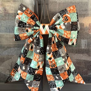 May include: A large decorative bow made of fabric featuring a patchwork pattern of Halloween-themed illustrations including pumpkins, ghosts, spider webs, and skulls in shades of orange, black, white, and mint green, with a central diamond-patterned knot.