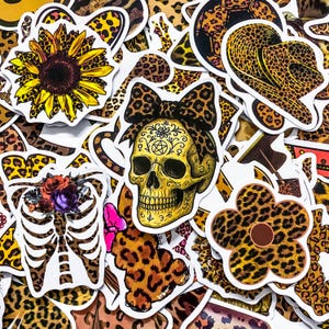 Peut inclure: Une collection d'autocollants colorés avec divers motifs. Comprend un tournesol, un crâne avec un nœud léopard, une cage thoracique avec des fleurs et des motifs léopard. Les autocollants sont sur fond blanc.