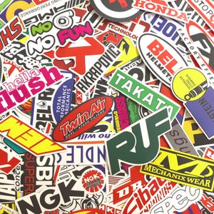 Può includere: Una collezione di adesivi colorati con vari loghi e testo. Gli adesivi presentano marchi come Honda, Takata e Twin Air, con una varietà di forme e dimensioni. Il design complessivo è un collage di grafica a tema automobilistico e da corsa.