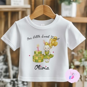 May include: White short-sleeved t-shirt featuring a graphic of a bunny holding balloons next to a cake with a number one candle. The text reads "One Little Sweet Twirl" and "Olivia".