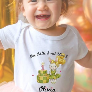 May include: White onesie with the text "One Little Sweet Twirl" and the name "Olivia". The onesie features a cartoon illustration of a bunny holding balloons and a birthday cake with a number one candle.