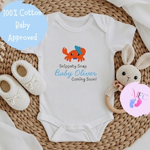 May include: White baby onesie with a crab graphic, the text "Snippet Snap Baby Oliver Coming Soon!" and the words "100% Cotton Baby Approved". Includes white baby shoes and a crochet bunny rattle.
