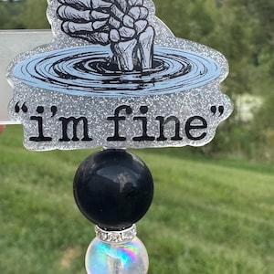 May include: A badge reel featuring a glittery acrylic topper with a skeleton hand giving a thumbs up from a blue water ripple graphic. The text "i'm fine" is printed in black below the graphic. A black bead and an iridescent bead are attached.