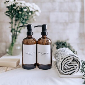 May include: Two brown glass bottles with black pumps labelled "HAND WASH" and "HAND LOTION" stand on a white surface. A rolled grey and white towel and a bar of soap are also present. The background includes white flowers and a white brick wall.