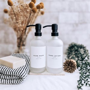 May include: Two frosted glass bottles with black pumps labelled "HAND WASH" and "HAND LOTION" sit on a stone base. A bar of soap rests on a black and white striped towel. Dried flowers and greenery complete the spa-like scene.