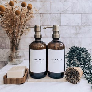 May include: Two amber glass bottles with metal pumps labeled "HAND WASH" and "DISH WASH". A wooden soap dish with a bar of soap, a small brush, and a vase of dried flowers are also present. The bottles sit on a white surface.