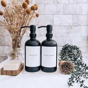 May include: Two black pump bottles labelled "SHAMPOO" and "CONDITIONER" with white labels. A wooden soap dish holds a bar of soap. A small brush and greenery are also present. The background features a white brick wall and a vase of dried flowers.