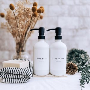 Op de afbeelding: Twee witte pompflessen met de labels "DISH WASH" en "HAND LOTION" met zwarte pompen. Een stuk zeep ligt op een zwart-wit gestreepte handdoek. Droogbloemen in een vaas staan op de achtergrond.