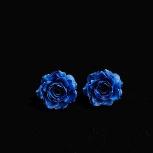 May include: Two vibrant blue rose-shaped earrings against a black background. The earrings have detailed petals and a glossy finish, creating a striking contrast. The floral design adds a touch of elegance.