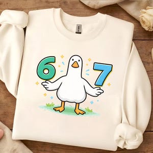 Puede incluir: Sudadera color crema con un diseño de pato de dibujos animados que sostiene los números 6 y 7. El pato es blanco con patas y pico naranjas, de pie sobre hierba verde. Los números son turquesa y azul, con destellos decorativos.