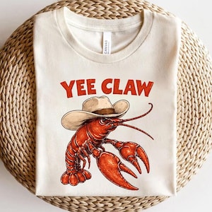 Peut inclure: T-shirt crème avec un dessin animé d'écrevisse portant un chapeau de cow-boy. L'écrevisse est rouge avec de grandes pinces et de longues antennes. Le texte "YEE CLAW" est imprimé en rouge au-dessus de l'écrevisse, dans un style vintage.