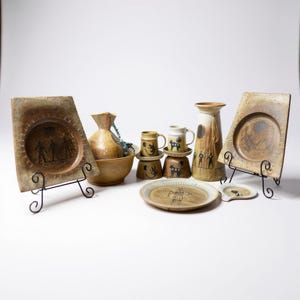 May include: A collection of handcrafted ceramic pottery with a rustic, earthy aesthetic. The assortment includes plates, mugs, a vase, and a decorative bowl, all featuring unique hand-painted designs in shades of brown and beige. The plates are displayed on black metal stands.