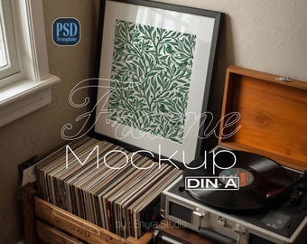 May include: A framed print with a green botanical pattern, a wooden crate filled with vinyl records, and a vintage record player. The image includes the text "Frame Mockup" and "by: Enyra Studio".