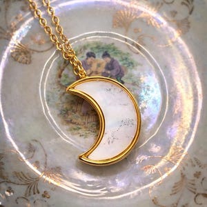 May include: A gold necklace with a crescent moon pendant. The pendant has a white, iridescent surface framed by a gold border. The necklace is displayed on a decorative plate with a painted scene and gold accents, creating an elegant aesthetic.