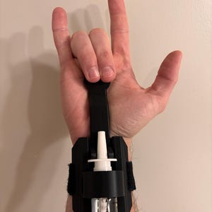 May include: A black wrist-mounted hand sanitizer dispenser with a clear bottle and white nozzle. The dispenser is secured with a black strap and velcro. The hand is making the 'rock on' hand gesture.