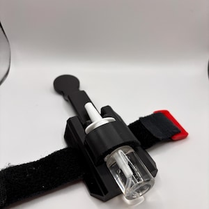 May include: A black nasal spray holder with a clear glass bottle and white nozzle. The holder is attached to a black adjustable strap with a red tab. The device is designed to hold a nasal spray bottle.