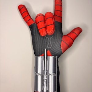 May include: A red and black fabric glove featuring a textured pattern and black web-like lines. The glove is positioned in a hand gesture with the index and pinky fingers extended. A silver metallic wrist attachment is secured to the forearm area.