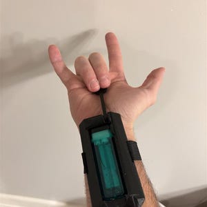 以下が含まれることがあります： 黒いリストマウントデバイス。緑と透明な円筒形のコンポーネントが付いています。デバイスは黒いストラップとコードで固定されています。デバイスは手首に装着されています。