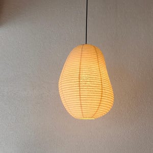 May include: A cream-colored, organic-shaped pendant lamp with a ribbed paper texture. The light fixture is suspended by a thin black cord from the ceiling, emitting a warm, soft glow through its translucent, layered material construction.