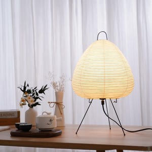 May include: A modern table lamp with a paper shade and black metal tripod base. The shade is a light cream color with horizontal lines. The lamp is on a wooden table, with a tea set and vases in the background.