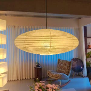 Op de afbeelding: Een grote, ovaalvormige hanglamp gemaakt van gebroken wit papier met horizontale geribbelde details. De lamp hangt aan een dun zwart snoer aan het plafond en verspreidt een zachte, warme gloed door het textuurrijke materiaal.