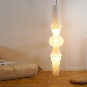 Könnte beinhalten: Eine hohe, cremefarbene Papierlaternen-Stehleuchte mit einem einzigartigen, gestapelten Design. Die Lampe hat einen zylindrischen oberen und unteren Teil mit einem kugelförmigen Mittelteil. Die Lampe ist beleuchtet und wirft einen warmen Schein. Die Lampe steht in einem Raum mit Holzboden.