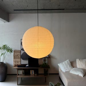 Pode incluir: Uma luminária pendente esférica de papel de grande porte com uma iluminação quente e brilhante. A cúpula apresenta uma textura delicada e nervurada com linhas horizontais finas na superfície, suspensa do teto por um fio preto fino.