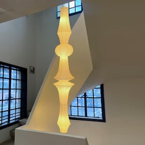 May include: A tall, cylindrical pendant lamp made of off-white paper with a visible horizontal ribbed texture. The lamp features a stacked, segmented design with varying widths, creating a series of rounded and tapered sections that emit a soft glow.