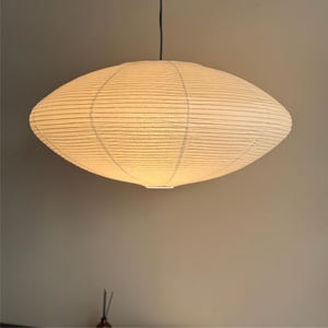 May include: A large, cream-colored paper pendant light fixture. The light is oval-shaped with a ribbed texture and is suspended from a black cord. The light is illuminated, casting a warm glow.
