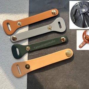 Może przedstawiać: Cztery skórzane osłony uchwytów w odcieniach pomarańczu, szarości, zieleni i beżu. Każda osłona ma prostokątny kształt z zaokrąglonym końcem i zapięciem na zatrzask ze złotym guzikiem. Tekst "Tailorstrapsstudio" jest wydrukowany na jednej z zielonych osłon.