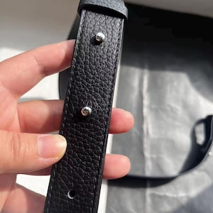 Op de afbeelding: Close-up van een zwarte leren riem met een getextureerd oppervlak. De riem heeft zilverkleurige klinknagels en een gesp. Het leer lijkt van hoge kwaliteit te zijn, met zichtbare nerf.