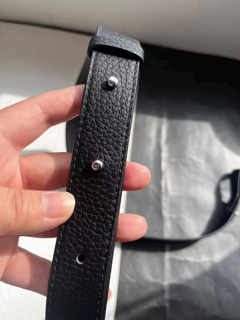 Op de afbeelding: Close-up van een zwarte leren riem met een getextureerd oppervlak. De riem heeft zilverkleurige klinknagels en een gesp. Het leer lijkt van hoge kwaliteit te zijn, met zichtbare nerf.