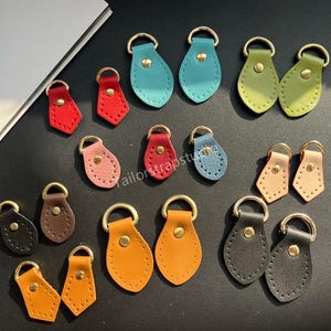 May include: A collection of teardrop-shaped leather tabs in various colors including red, blue, green, yellow, brown, black, and beige. Each tab features a gold-toned metal D-ring at the top, a central gold-toned rivet, and decorative stitching.