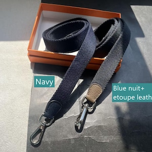 May include: Two navy blue and blue-gray shoulder straps with silver-tone clasps. One strap is solid navy, the other is blue-gray with a tan leather accent. The straps are displayed in an orange box.