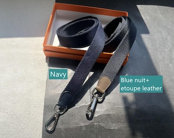 May include: Two navy blue and blue-gray shoulder straps with silver-tone clasps. One strap is solid navy, the other is blue-gray with a tan leather accent. The straps are displayed in an orange box.