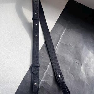 Op de afbeelding: Een zwarte leren tasriem met zilverkleurige metalen hardware. De riem heeft een getextureerd oppervlak en is verstelbaar in lengte. Het ontwerp omvat een gesp en verschillende klinknagels. De achtergrond is een mix van zwarte en witte geometrische vormen.