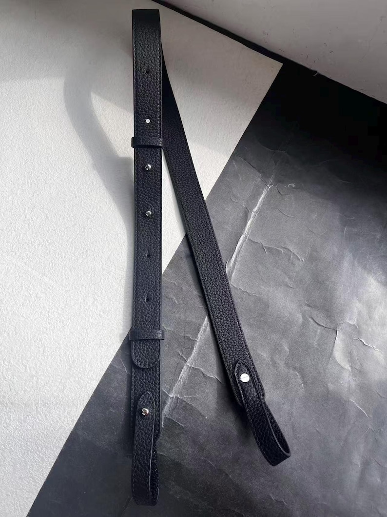 Op de afbeelding: Een zwarte leren tasriem met zilverkleurige metalen hardware. De riem heeft een getextureerd oppervlak en is verstelbaar in lengte. Het ontwerp omvat een gesp en verschillende klinknagels. De achtergrond is een mix van zwarte en witte geometrische vormen.