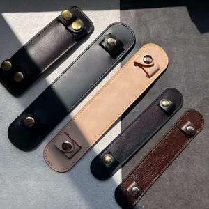 May include: A collection of leather pen holders in various colors, including black, brown, and beige. Each holder features a snap closure with gold-colored hardware. The holders are rectangular and designed to hold a single pen.
