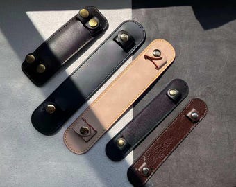 Op de afbeelding: Een verzameling leren penhouders in verschillende kleuren, waaronder zwart, bruin en beige. Elke houder heeft een druksluiting met goudkleurige hardware. De rechthoekige houders zijn ontworpen om één pen te bevatten.