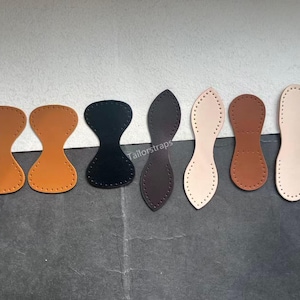 May include: A collection of leather bag bases in various colors, including tan, black, and brown. Each piece is shaped for bag construction, with pre-punched holes for stitching. The pieces are arranged on a neutral surface.
