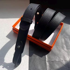May include: A black leather belt with a textured finish, displayed in an orange box with a white interior. The belt is folded and features a buckle and a loop. The background is a mix of grey and white.