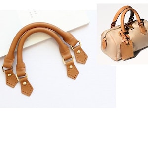 May include: Two sets of light brown leather bag handles with gold-colored hardware. One set is lying flat, and the other is attached to a small, beige handbag with matching leather accents and a luggage tag.