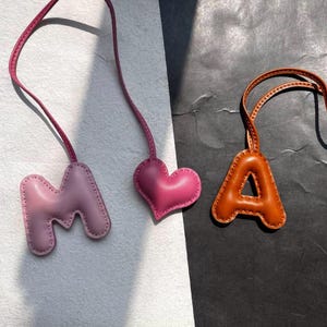 Puede incluir: Tres colgantes de cuero: una letra "M" rosa claro, un corazón rosa y una letra "A" marrón. Cada colgante tiene una correa a juego para colgar. Los colgantes están cosidos alrededor de los bordes.