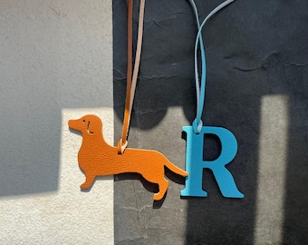 Puede incluir: Un charm de perro salchicha de cuero naranja y un charm azul con la letra "R", ambos con cordones colgantes. El charm del perro tiene textura. El fondo es una mezcla de blanco y gris oscuro.