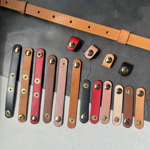 May include: A collection of leather strap accessories in various colors, including black, brown, red, and pink, with gold-tone metal hardware. A tan leather strap is visible at the top. The accessories are designed for use with bags or other leather goods.